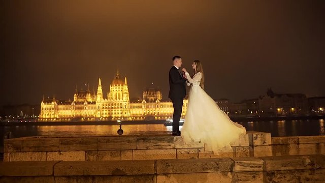 young loving couple in wedding.  are standing cuddling on the background of the old night city.  illuminated Parliament building, Budapest Hungary.  Europe.  castle lights in the night. Slow motion