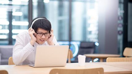 Young Asian man wearing headphones listening to music while working with laptop computer at home office. Relaxing at work concept