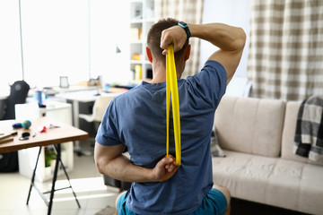 Close-up of man doing exercise for back using fitness rubber band. Home workout on quarantine period. Cozy living room interior. Strong and fit body. Sport and active lifestyle concept