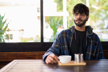 Fototapeta na wymiar Young handsome bearded Indian hipster man thinking at the coffee shop