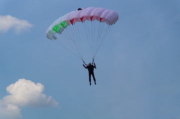 A skydiver in black outfit and black glasses flies across the sky with clouds on a white-red-green parachute