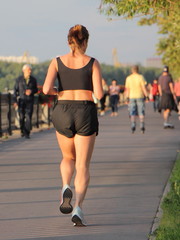 One European athletic young woman in light summer sportswear runs along an asphalt alley at a training session after a working day, rear view