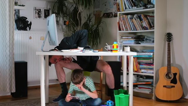 Father is doing business chores in the home office. He sits under the table to his son. Combining work and pedagogy in self-expression.