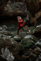 Attractive blonde curly young woman jumping from stone to stone, wearing red authentic poncho with ethnic pattern, in the beautiful stony cave with the creek 