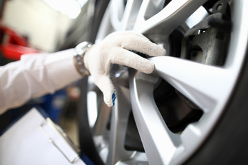 Close-up gloved hand touches the cap car wheel. An auto mechanic holds in one hand a clipboard with technical information. Braking distance calculation. Professional forensic accident