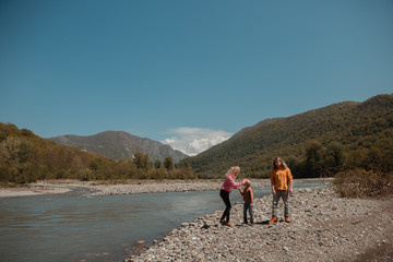 Fototapeta premium Beautiful young family, dad, mother and little son, having fun on the beach of mountain river near green summer forest with great view in the bright sunny day, blue sky backward