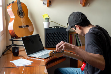 Young man in cap tuning and learn electric guitar using a laptop at home. Musician playing guitar...