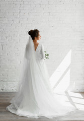 beautiful young bride with a bouquet of beautiful flowers posing against a white studio brick wall,gorgeous wedding dress of a pretty bride,fashionable wedding dress,floristry of a wedding bouquet