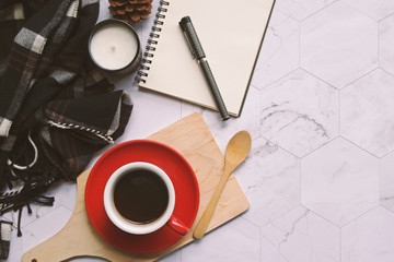 The top view of the red coffee cup with black coffee inside it on the tray, and have a wooden spoon beside the cup.Near the cup of coffee is a black scarf, candle and a notebook on the table.
