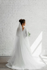 beautiful young bride with a bouquet of beautiful flowers posing against a white studio brick wall,gorgeous wedding dress of a pretty bride,fashionable wedding dress,floristry of a wedding bouquet