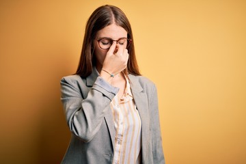 Young beautiful redhead woman wearing jacket and glasses over isolated yellow background tired rubbing nose and eyes feeling fatigue and headache. Stress and frustration concept.