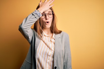 Young beautiful redhead woman wearing jacket and glasses over isolated yellow background surprised...