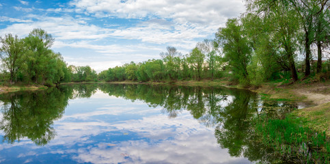 lake or creek surrounded by trees, with crystal clear and calm water, which reflects the sky and clouds on a sunny day