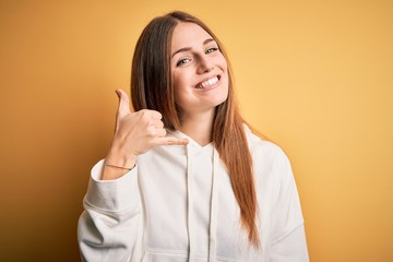 Young beautiful redhead sporty woman wearing sweatshirt over isolated yellow background smiling doing phone gesture with hand and fingers like talking on the telephone. Communicating concepts.