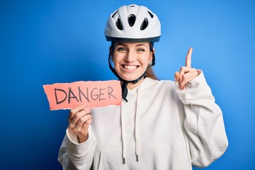 Young beautiful redhead cyclist woman wearing bike helmet holding danger paper message surprised with an idea or question pointing finger with happy face, number one