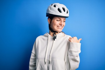 Young beautiful redhead cyclist woman wearing bike helmet over isolated blue background smiling with happy face looking and pointing to the side with thumb up.