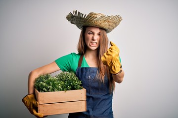 Young beautiful redhead farmer woman wearing apron and hat holding box with plants annoyed and frustrated shouting with anger, crazy and yelling with raised hand, anger concept