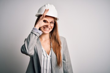 Young beautiful redhead architect woman wearing security helmet over white background doing ok gesture with hand smiling, eye looking through fingers with happy face.
