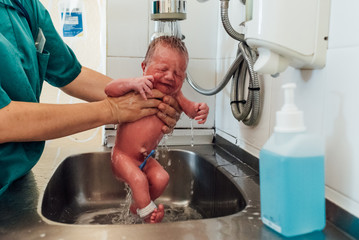 Nurse washes a newborn baby under the tap in the hospital. She holds the child with her 2 hands...