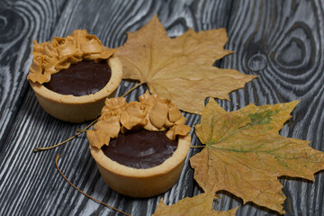 Tartlets with chocolate ganache. On autumn maple leaves. Decorated with oil cream flowers. The cream has a caramel color. On brushed pine boards painted black.