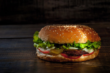 Burger on a dark wooden board. Cheeseburger with cutlet and herbs on black background. Food with contrasting dramatic light. Bun closeup.