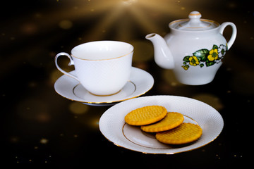 The tea set, consisting of a tea cup with a saucer, a kettle and a plate of biscuits, is located on a black background. Sunlight shines from the top.