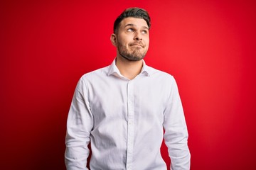 Young business man with blue eyes wearing elegant shirt standing over red isolated background smiling looking to the side and staring away thinking.