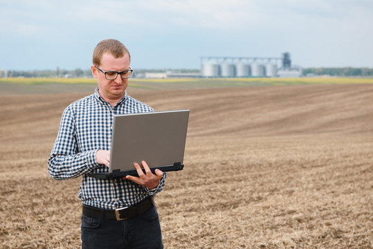 Happy Farmer With Laptop Standing In Wheat Field In Front Of Grain Silo