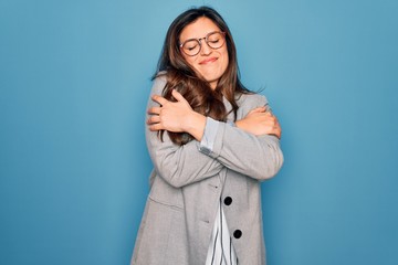 Young hispanic business woman wearing glasses standing over blue isolated background Hugging oneself happy and positive, smiling confident. Self love and self care