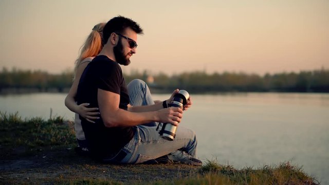 Couple Sitting By The River With Cups Of Tea. Couple Relaxing End Watching On Sunset. Couple Is Looking At Red Sunset.
