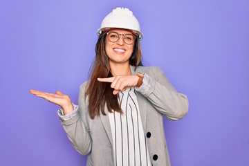 Professional woman engineer wearing industrial safety helmet over pruple background amazed and smiling to the camera while presenting with hand and pointing with finger.