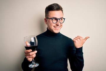 Young handsome caucasian man drinking an alcoholic glass of red wine over isolated background pointing and showing with thumb up to the side with happy face smiling