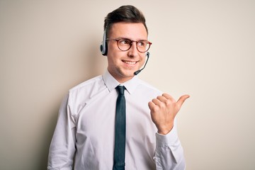 Young handsome caucasian business man wearing call center headset at customer service smiling with happy face looking and pointing to the side with thumb up.