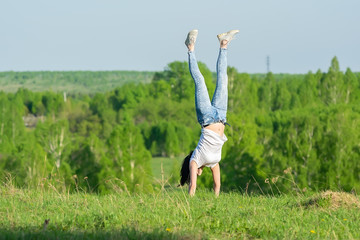 a sporty looking girl stands on her hands upside down on a hillock against the background of a forest near the village