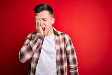 Young handsome caucasian man wearing casual modern shirt over red isolated background Yawning tired covering half face, eye and mouth with hand. Face hurts in pain.