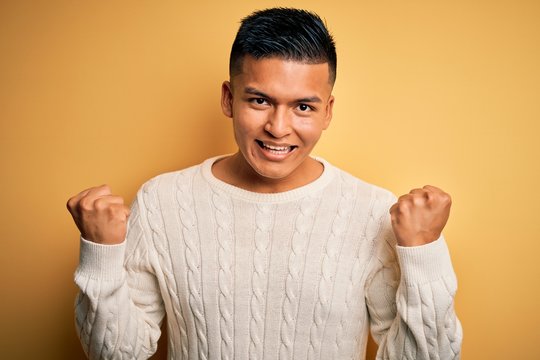 Young handsome latin man wearing white casual sweater over yellow background celebrating surprised and amazed for success with arms raised and open eyes. Winner concept.