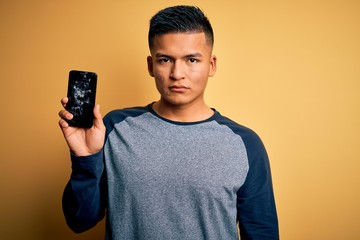 Young handsome latin man holding broken smartphone showing craked screen with a confident...