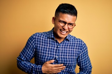 Young handsome latin man wearing casual shirt and glasses over yellow background smiling and laughing hard out loud because funny crazy joke with hands on body.