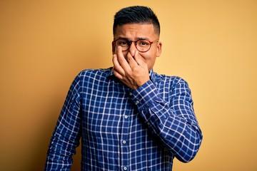 Obraz premium Young handsome latin man wearing casual shirt and glasses over yellow background smelling something stinky and disgusting, intolerable smell, holding breath with fingers on nose. Bad smell
