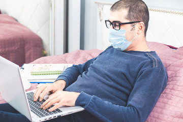 Fototapeta na wymiar Man in protective mask working from home during quarantine time at coronavirus Covid-19 outbreak. He is sitting on the bed with laptop.