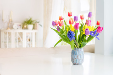 Fresh spring colorful bouquet of tulips, daffodils, irises in vase standing on white marble table with light classic design living room background. Festive flowers for gift. Mockup for greeting card.