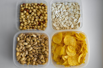 Top view of various delicious crispy snacks such as pistachio nuts, salted pumpkin seed, fried hazelnuts and potato chips which are stored in plastic transparent containers