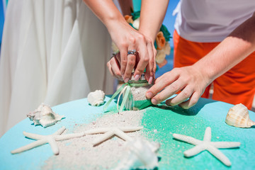 
Bride and groom pouring colorful different colored sands into the crystal vase close up during symbolic nautical decor destination wedding marriage ceremony on sandy beach in front of the ocean