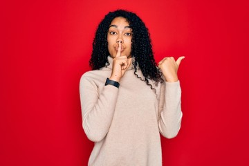 Young african american curly woman wearing casual turtleneck sweater over red background asking to be quiet with finger on lips pointing with hand to the side. Silence and secret concept.