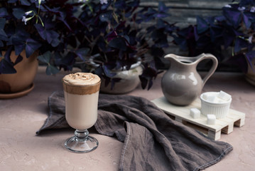 A popular Korean drink during self-isolation is Dalgona coffee. A delicious modern cold beverage with sugar, creamer and napkin is on the table. On a background is potted plants. Vintage still life.