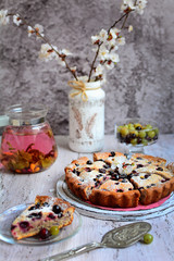 View of homemade pie with berries, herbal tea in a glass teapot, a piece of cake on a plate and flowering apricot branches in a vase on a wooden table. Home Relax