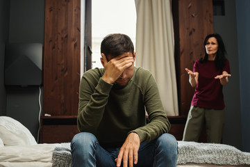 Couple with age gap or mother and adult son arguing in bedroom. Anxious middle aged woman blaming depressed young man sitting on bed and covering face with hand