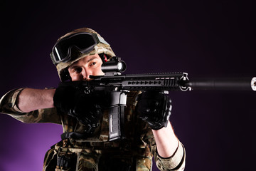 Fototapeta na wymiar Soldier with gun is looking through the scope on violet background. Concept of war. Veterans, comrades, soldiers. Man in uniform.