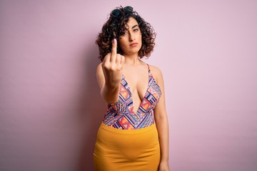 Young beautiful arab woman on vacation wearing swimsuit and sunglasses over pink background Showing middle finger, impolite and rude fuck off expression