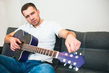 Young man sitting in room during quarantine. Guy adjusting strings for better playing on acoustic guitar. Solo musician learning to play well. © Vlad